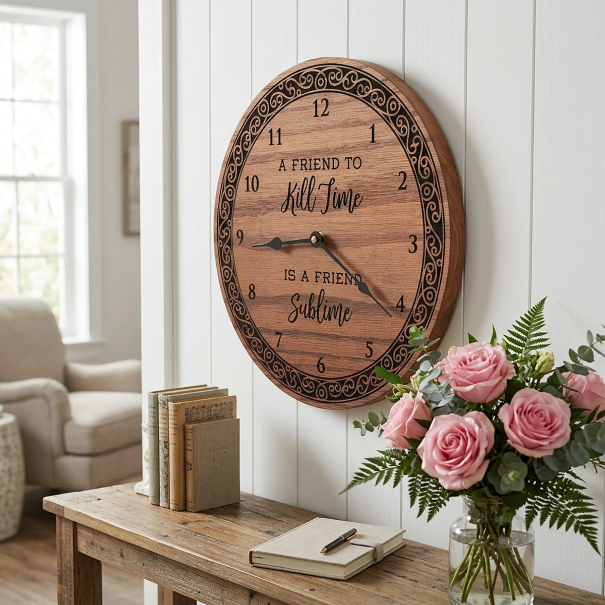 An oval wooden wall clock with engraved black numerals and the phrase 'A FRIEND TO Kill Time IS A FRIEND Sublime' hangs on a white paneled wall above a rustic wooden console table. The table holds pink roses in a glass vase, a notebook, and a stack of books, with a softly blurred living room in the background.