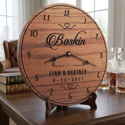 A personalized wooden clock styled like a wine barrel, standing on a small wooden easel. The clock features black engraved numbers, hands, and text reading 'Baskin', 'FINN & HEATHER', and '8.