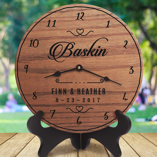 A round wooden clock with engraved black numerals, hands, and personalized text reading 'Baskin' and 'Finn & Heather 8-23-2017,' displayed on a black stand against a blurred outdoor park background.