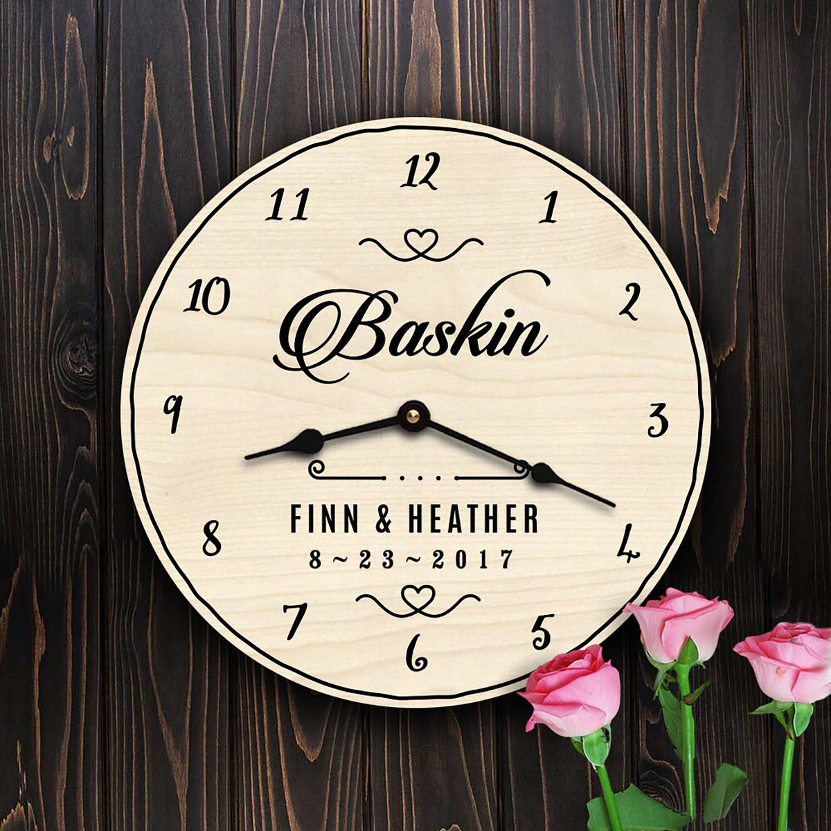 A personalized round wall clock with a light wood grain face and black numerals, hands, and custom text reading 'Baskin' and 'FINN & HEATHER 8~23~2017', shown against a dark wood plank background with pink roses at the bottom right.