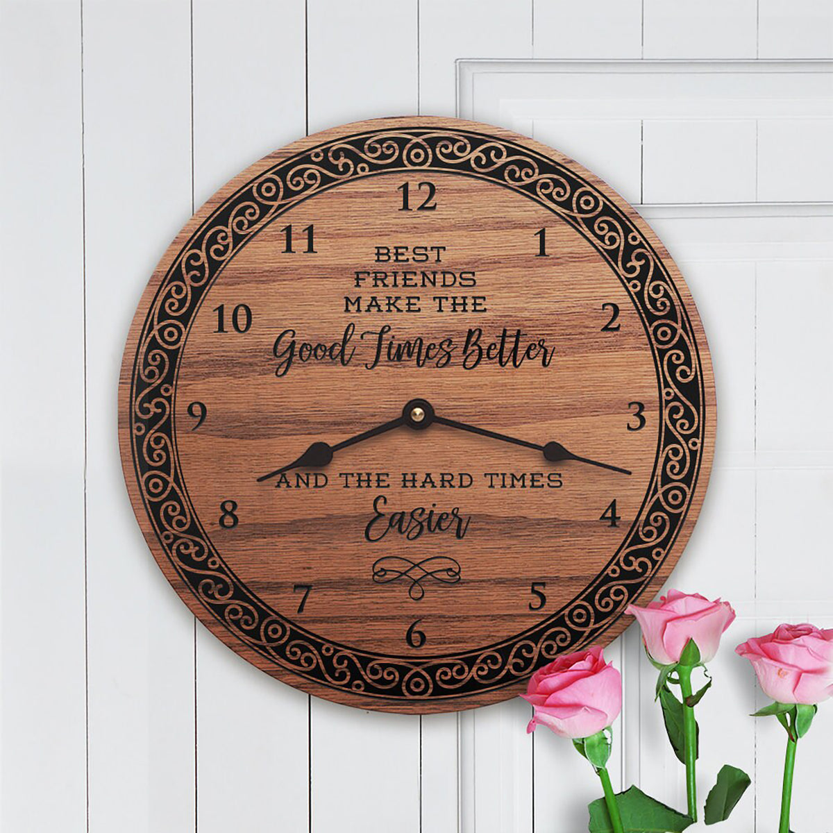 A round wooden wall clock with a decorative black scroll border and engraved text reading 'Best Friends Make the Good Times Better and the Hard Times Easier.' The clock is shown against a white paneled wall with pink roses at the bottom right.