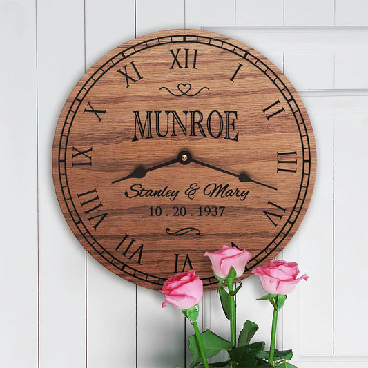 Personalized round wooden wall clock with black Roman numerals and hands, engraved with the name Munroe, shown against a white paneled wall with pink roses at the bottom.